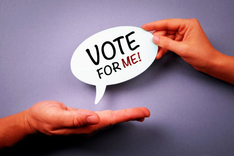 Hand holding a speech bubble that says 'VOTE FOR ME!' above another open hand.